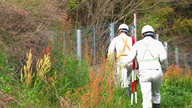 測量機材を担いで現場へ向かう2人